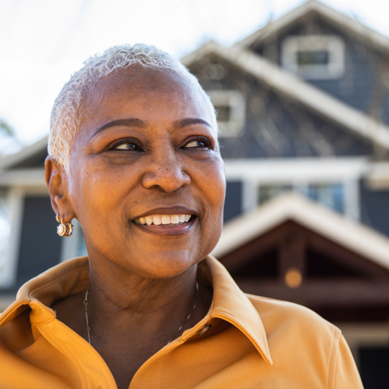 A middle aged woman smiling in front of her home knowing her retirement goals are protected.