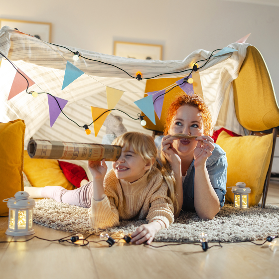 Woman and child smiling inside a blanket fort with string lights.