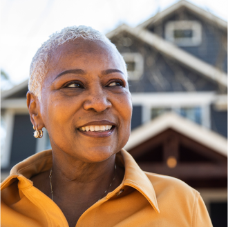 A middle aged woman smiling in front of her home knowing her retirement goals are protected.
